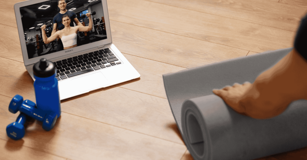 A person unrolls a yoga mat in front of a laptop showing a virtual workout session with fitness instructors.